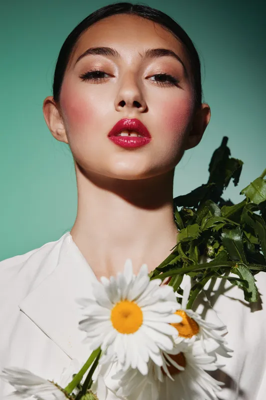 Close-up of a young woman with glossy red lips, shimmering eyeshadow, and blush holding white daisies. Concept of beauty creativity fashion with natural floral elements.