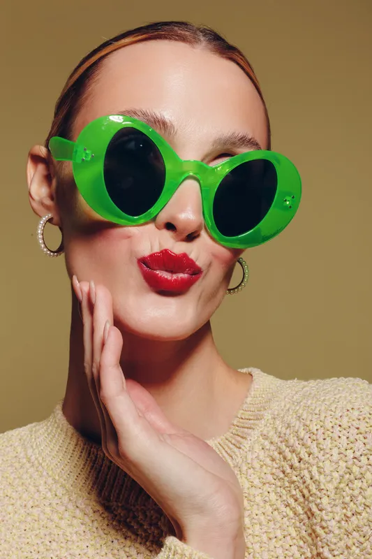 Woman with green sunglasses and red lipstick poses with hand near face on beige background. Fashion portrait with bright makeup and stylish accessories in studio lighting.