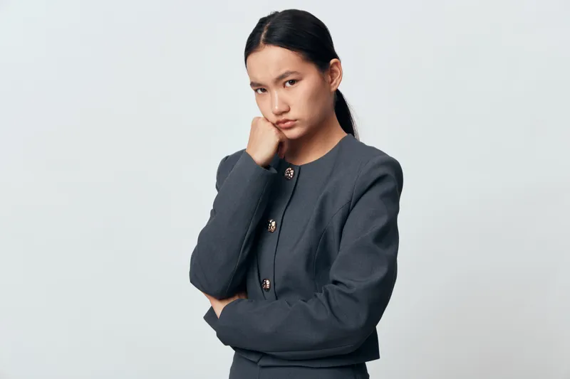 Young woman with dark hair in gray business suit posing with thoughtful expression and hand on chin against white background, studio portrait for professional concept.