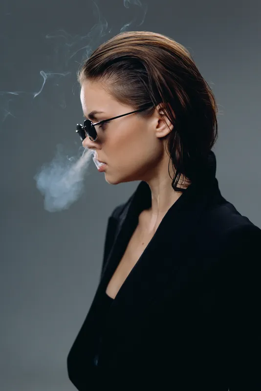 Profile of a smoking woman in sunglasses wearing dark stylish blazer with smoky background and serious expression