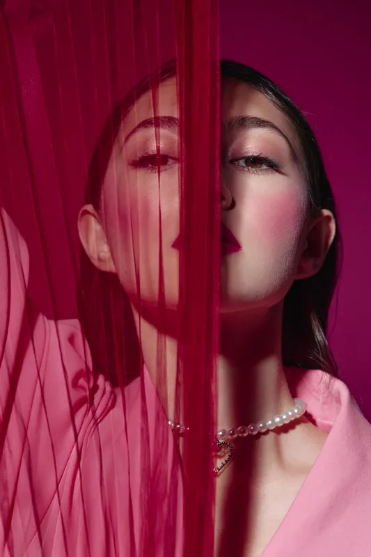 Creative beauty portrait of a young woman behind sheer pink fabric wearing pearl necklace and soft pink jacket on magenta background. Fashion and artistic concept.