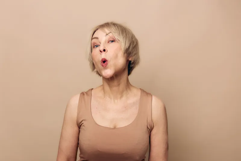 Mature woman with short blonde hair wearing beige tank top makes surprised expression with pursed lips against plain beige background. Concept of emotions, age, and natural beauty.