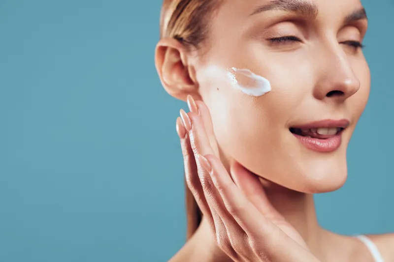Young woman applies moisturizing cream on her cheek with closed eyes enjoying skincare routine isolated on blue background natural beauty healthy skin care concept.