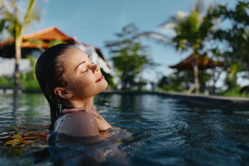 A woman enjoys warm sunlight as she relaxes in a bright outdoor pool, water caressing her shoulders. Calm, refreshing atmosphere, leisure and wellness tied to summer self-care and tranquil moments.