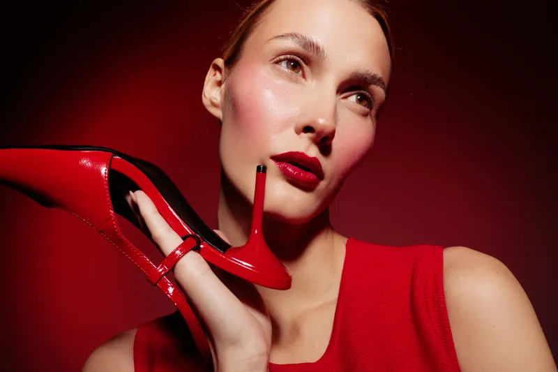 Close-up of a stylish woman in red dress holding a red high heel shoe near her face. Glossy makeup and shiny skin create a glamorous and elegant fashion concept on red background.