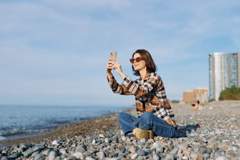 Woman on a pebble beach takes a selfie with a smartphone, wearing plaid shirt, jeans and sunglasses, enjoying sunny seaside scenery and casual outdoor leisure, travel lifestyle moment by the water