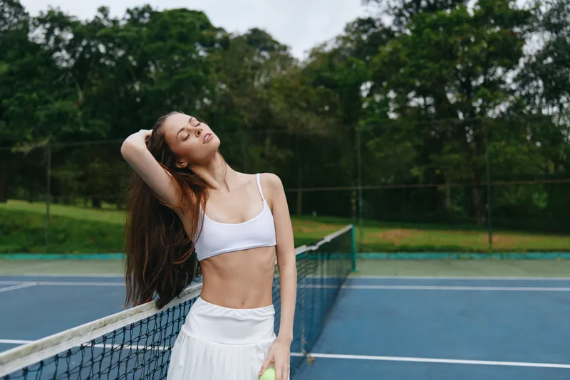 Young athletic woman in white sportswear enjoying a moment on a tennis court with lush greenery, expressing joy and confidence