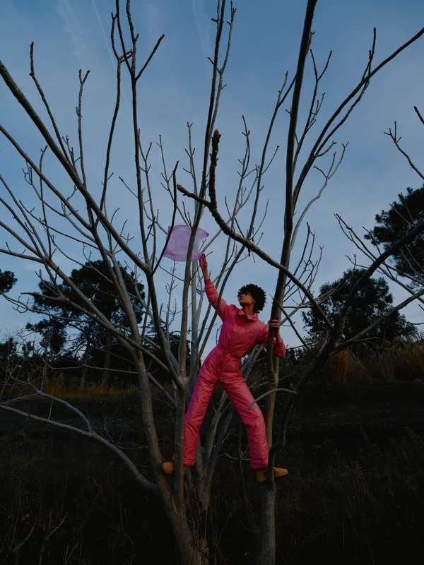 Person wearing a pink jumpsuit climbs a bare tree outdoors reaching branches with effort in a natural setting under a cloudy sky and sunlight hint capturing a moment of physical challenge and outdoor