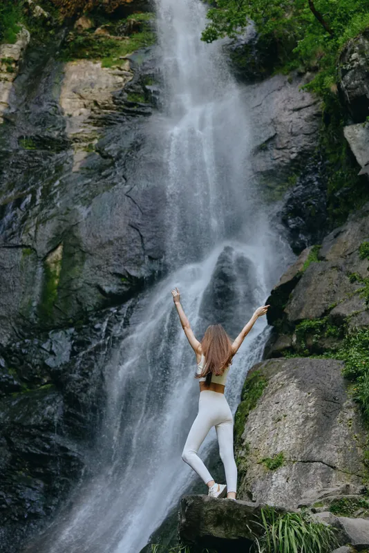 Woman standing with raised arms in front of tall natural waterfall surrounded by green foliage and rocky cliffs, expressing freedom and connection with nature.