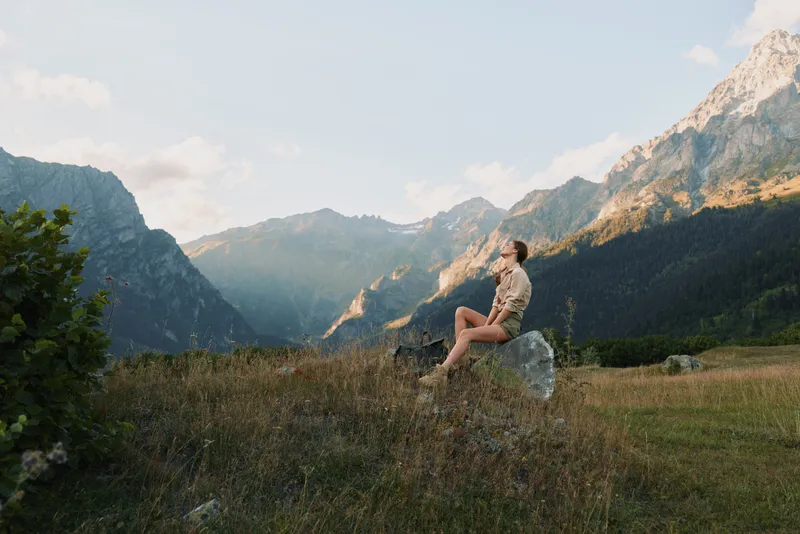 Woman enjoying outdoor relaxation in a meadow beneath towering mountains, sitting on a rock, gazing at scenery, embodying a solo traveler mood and peaceful nature