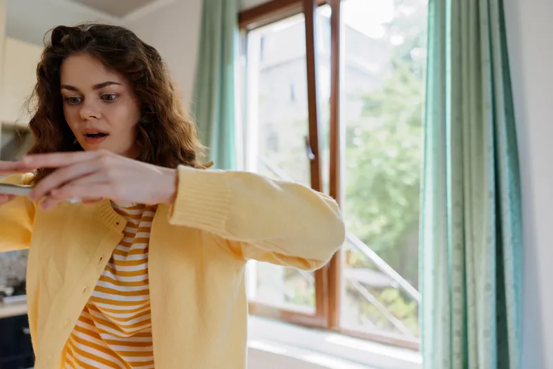 A young woman with curly hair wearing a yellow cardigan and striped top stretches her arms in a bright home setting, reacting with surprise in a casual morning moment.