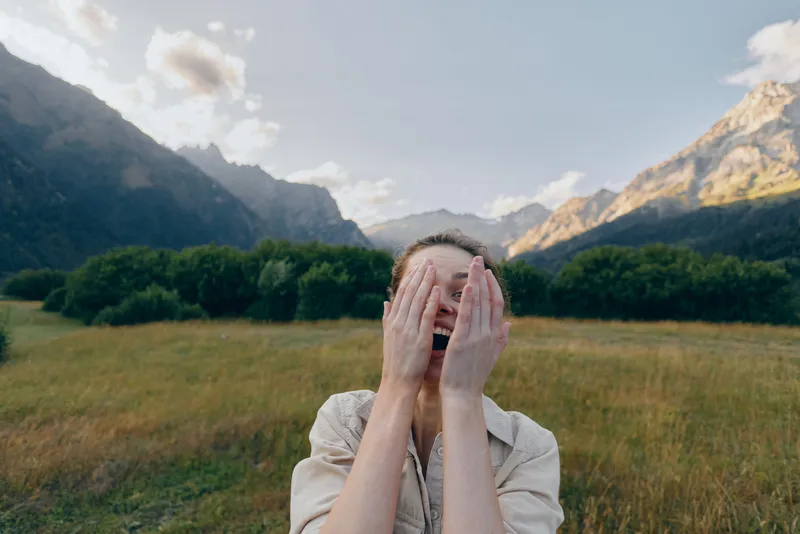 Young woman in a sunlit valley covers her face with hands, surprised and playful, green meadow and distant mountains in the background