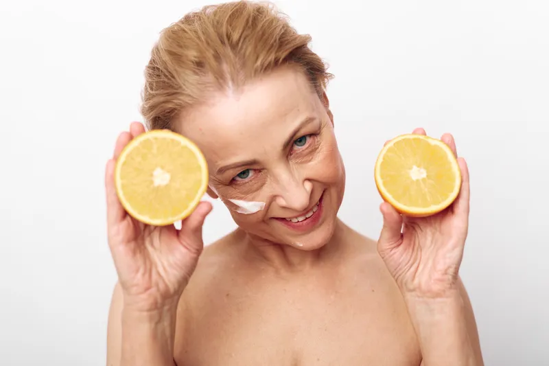 Mature woman with healthy skin holds two halves of fresh orange near face, smiling gently. Cream is applied on cheek, concept for natural skincare, wellness, and vitamin c benefits.