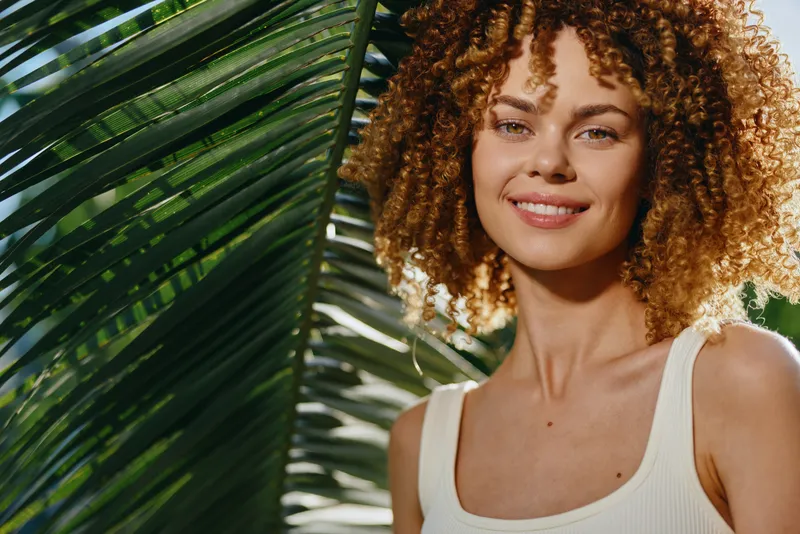 smiling woman with curly hair standing near tropical palm leaves, wearing white tank top, natural sunlight illuminating her face outdoors, warm summer vibe