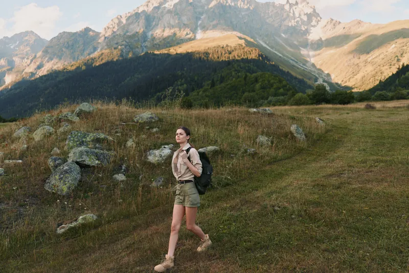 Woman hiking through a mountain meadow with a backpack, exploring nature on a dirt trail, showcasing an active outdoors lifestyle, calm adventurous mood in a wide open alpine landscape