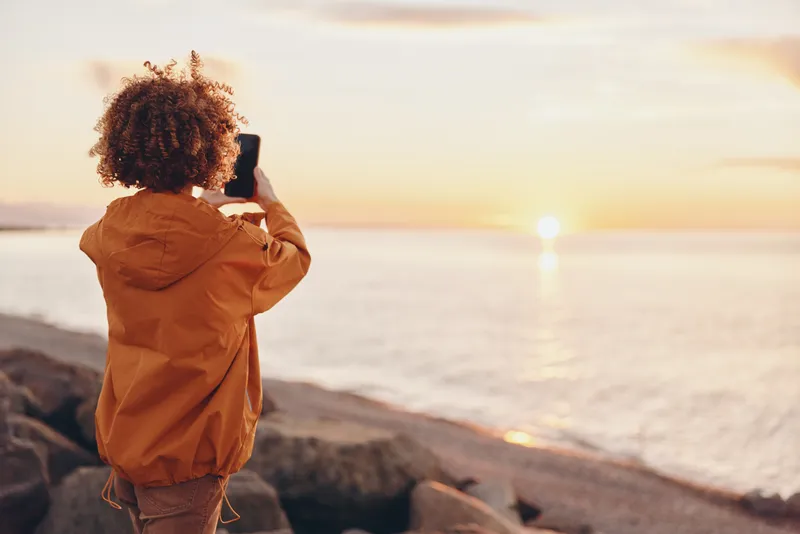 Woman lifestyle, back view, wearing orange jacket, stands on rocky beach by ocean at sunset, gazing at horizon, calm nature travel, peaceful solitude, soft atmosphere