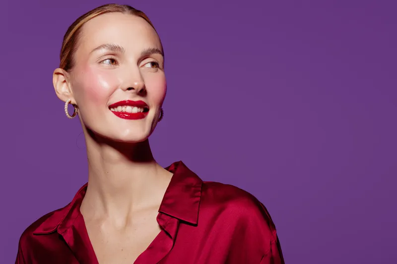 Smiling woman with red lipstick and gold hoop earrings wears red satin blouse against purple background. Concept of beauty, fashion, elegance, and confident female portrait.