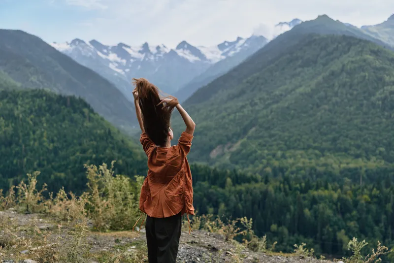Woman standing on a hillside overlooking a mountain valley, wearing a brown top, arms raised, enjoying the view of forested slopes and distant snow capped peaks.