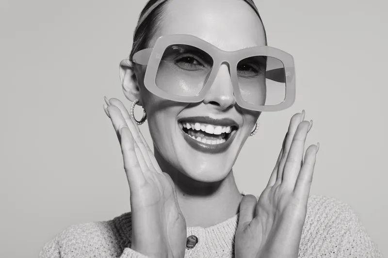Smiling white woman with light skin wearing large sunglasses and sweater shows happy expression with hands near face on plain background.