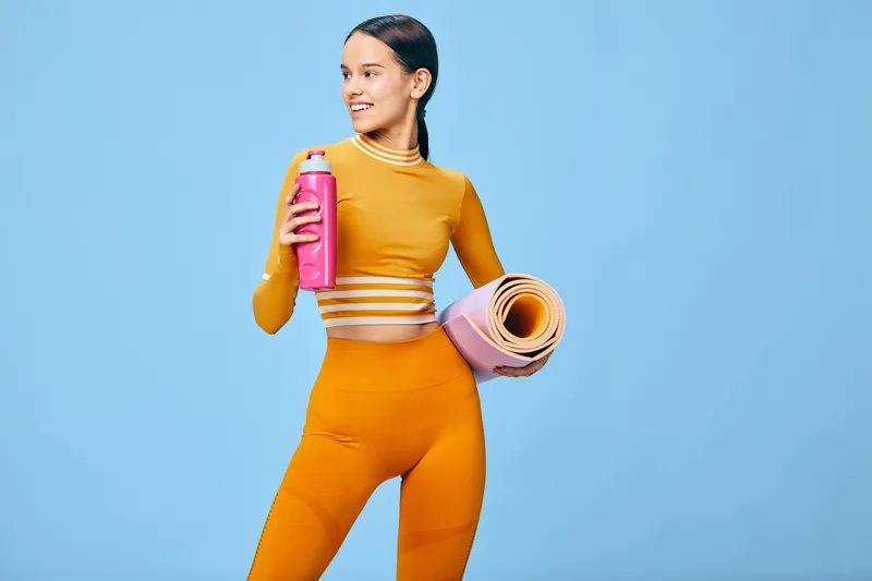 Woman fitness athlete in orange sportswear stands with a hydration bottle and rolled yoga mat against a blue studio backdrop, ready for gym workout and active lifestyle.