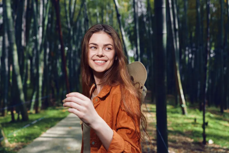 Woman with skateboard and smile in park on tree-lined path, portrait of young adult enjoying outdoors in orange jacket, carefree motion and natural expression during sunny walk.
