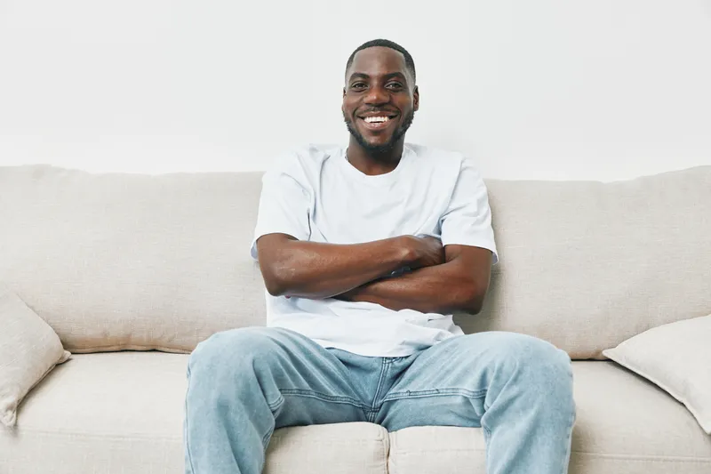 Cheerful young African American man in casual attire sitting on a sofa, smiling with a relaxed demeanor, conveying a sense of comfort and happiness