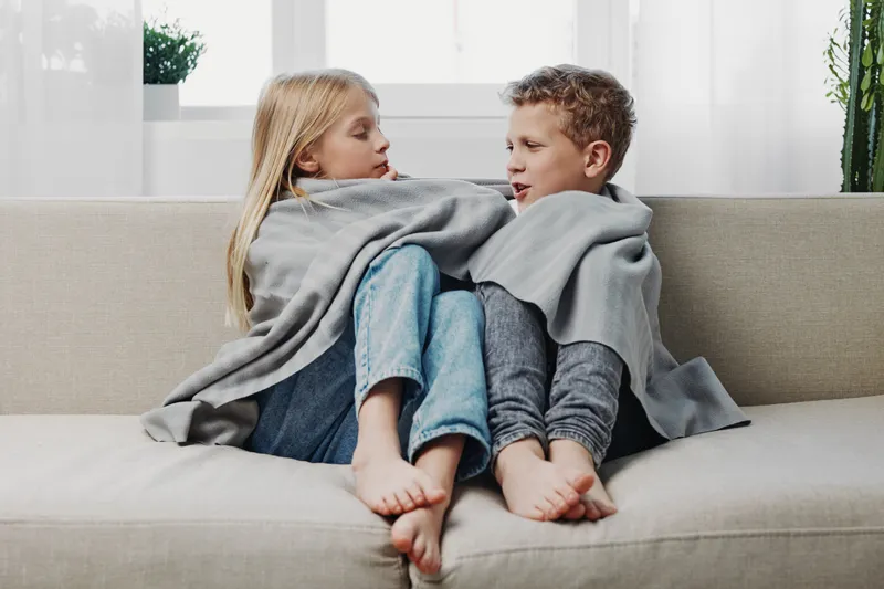 Two children wrapped in a gray blanket sit barefoot on a beige couch in a bright living room, sharing a quiet moment together indoors.