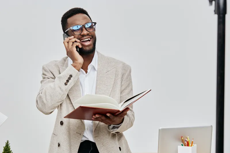 Happy smiling man wearing beige blazer and white shirt talking on mobile phone holding open notebook in office setting with laptop and colorful stationery on desk, real people lifestyle concept