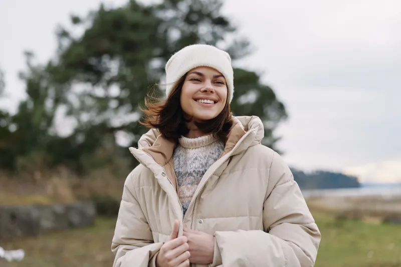 Woman outdoors wearing a beige puffer coat and white knit beanie, smiling on a windy day with trees and a lake in the distance, peaceful and warm.