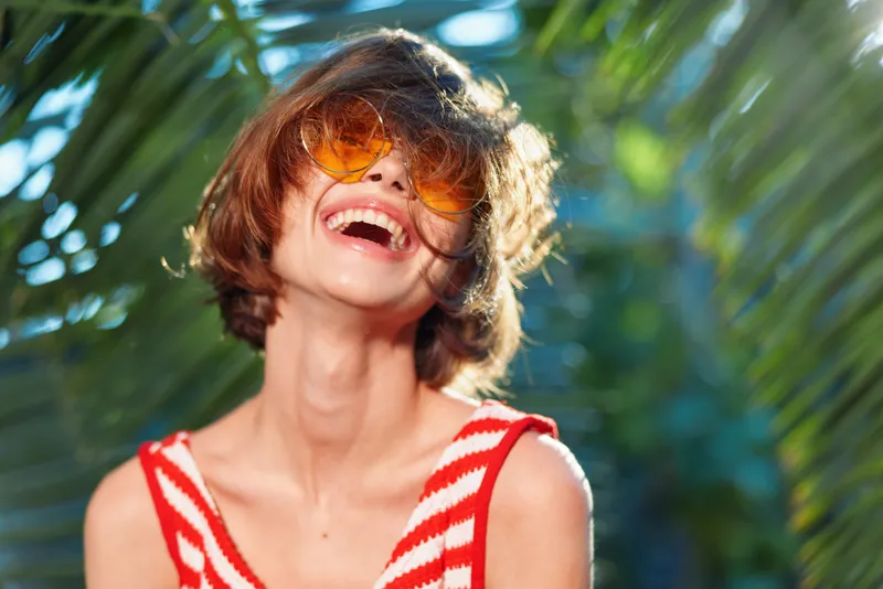 A cheerful young person wearing orange sunglasses and a striped red and white top, laughing outdoors among tropical plants, exuding carefree summer vibes and playful energy.