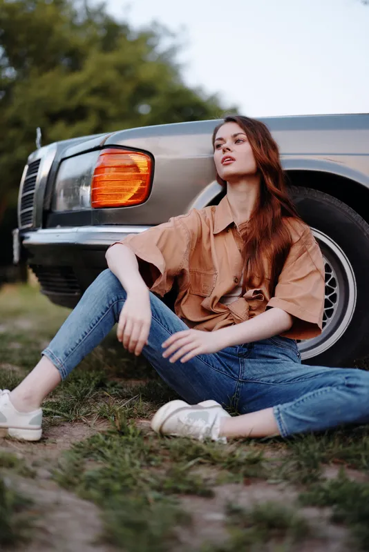Young woman in casual outfit sitting beside classic car in nature, showcasing relaxed summer vibes and modern fashion trends with a nostalgic touch.