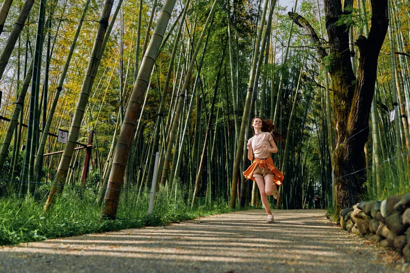 Woman running along a gravel path through dense bamboo forest in summer, flowing dress and motion, outdoor trail jog with sunlight filtering through tall green trees and foliage