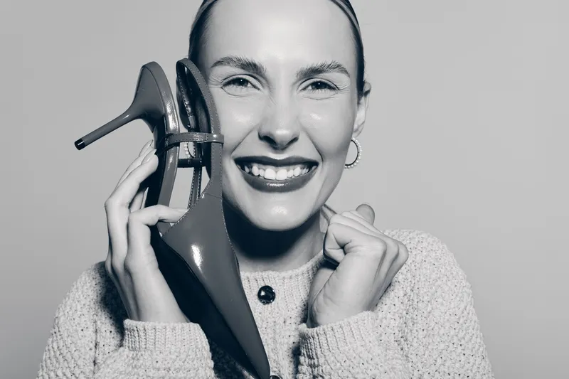 Happy smiling Caucasian woman with light skin holding high heel shoes close to her face in monochrome studio portrait.