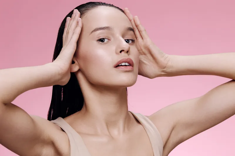 Young woman with wet hair expressing tranquility, wearing a beige top against a soft pink background, showing beauty and skin care concept