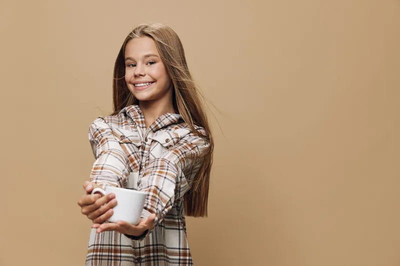 Teenage girl smiling and holding white cup for drink in casual plaid shirt with long hair on beige background for lifestyle and happiness.