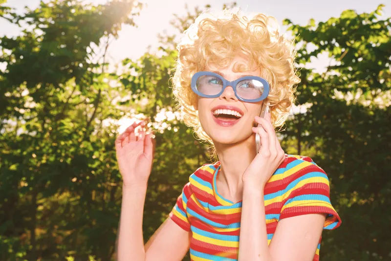 A cheerful woman in a striped shirt and oversized blue glasses enjoys a sunny day outdoors, radiating retro charm. Bright colors and natural light emphasize playful lifestyle.