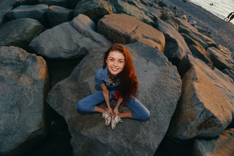 Lifestyle woman in modern boho-western denim street style sitting on rocks, smiling warmly at sunset with film color tones and natural outdoor beach setting.