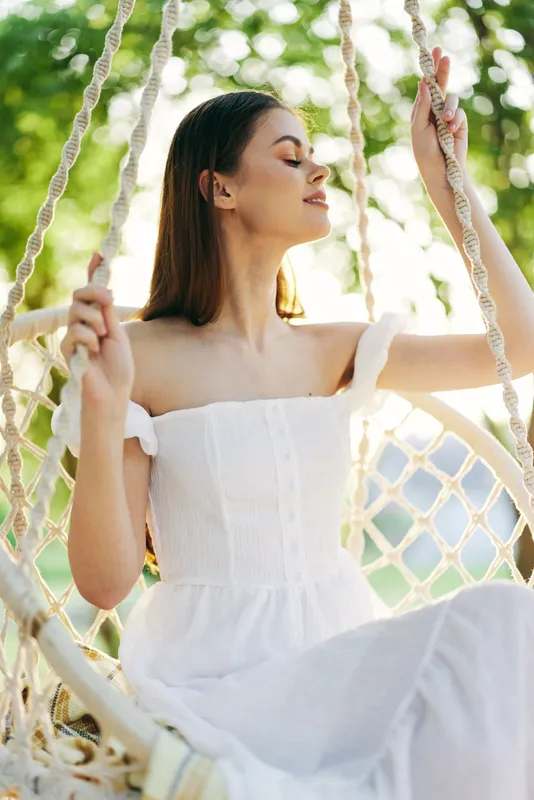 A serene young woman in a white dress enjoying a sunny day on a swing, surrounded by greenery, evoking feelings of relaxation and peace