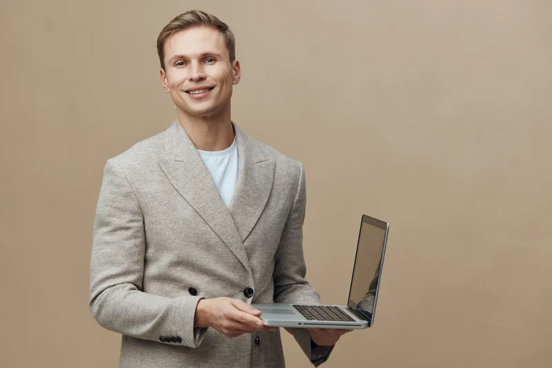 Smiling man in business casual outfit holding a laptop computer facing camera on beige background, studio portrait with confident expression. Professional, technology, modern, casual, cheerful, workpl