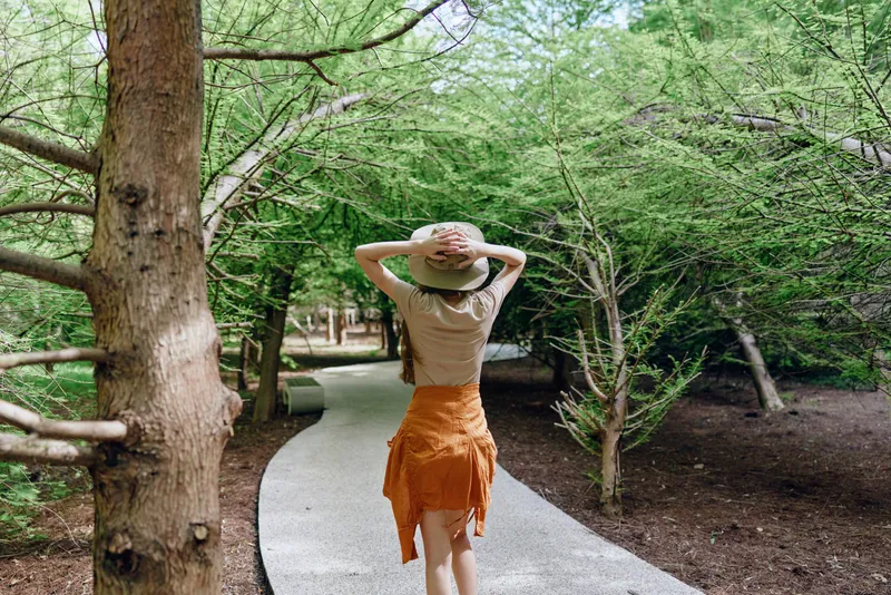 Woman walking on winding path through trees in a serene forest park, wearing a summer hat and orange skirt, hands on hat, exploring nature and enjoying a peaceful outdoor stroll.