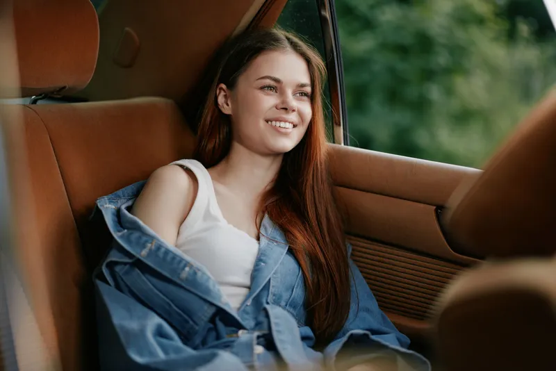 Young woman with long hair smiling in a car, wearing a denim jacket, reflecting ease and freedom in a lush green background. Seasonal vibe of summer.