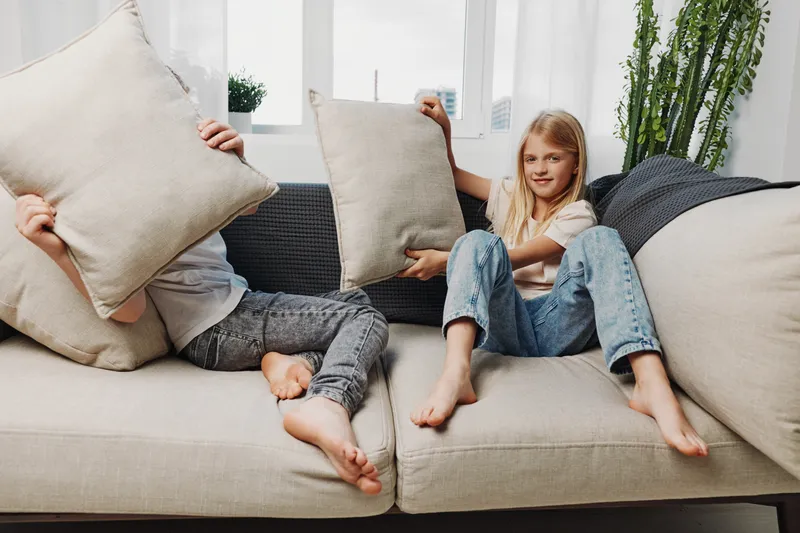 Two barefoot children having fun with pillows on a cozy sofa in a bright living room, enjoying playful moments and sibling interaction at home.