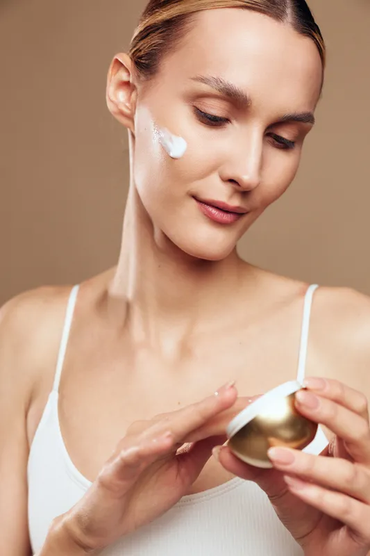 Young woman applies facial cream on cheek using fingers, demonstrates skincare routine with gentle expression, wearing white tank top, isolated on beige background.
