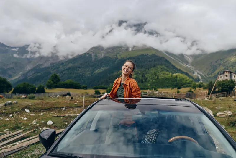 woman car mountains roadtrip countryside sunroof smile travel adventure cloudy sky authentic inclusivity joyful solo traveler enjoying nature and wide landscape