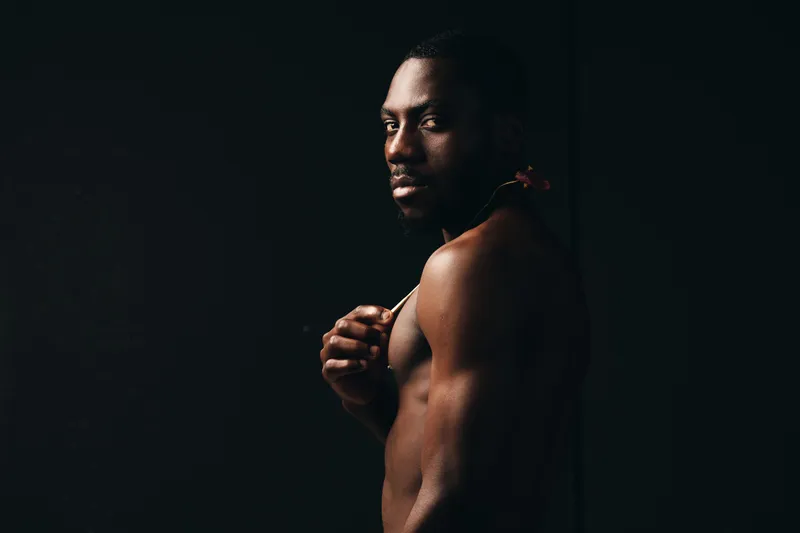 African man with dark skin posing shirtless in creative conceptual visual style, showing muscular body and serious expression on black background in studio lighting portrait.