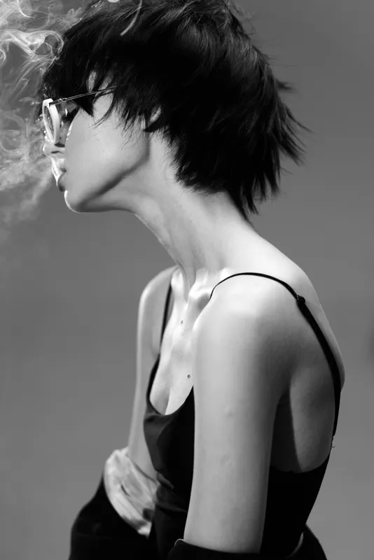 Black and white portrait of a young woman with short hair and glasses exhaling smoke in a stylish minimalist setting.