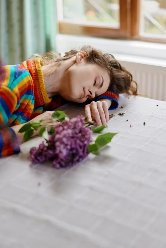 Calm woman indoors resting on a table with flowers, colorful striped sweater, soft daylight, peaceful mood, natural tones, intimate home moment, relaxed posture