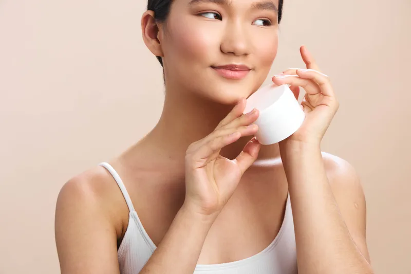 asian woman in a soft studio lights up with a gentle smile as she holds a skincare jar, presenting a calm, healthy lifestyle and a luminous complexion in a minimal setting.