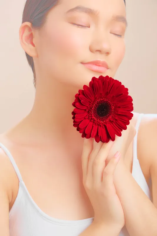 asian woman holds a vivid red flower near her face in a soft studio portrait. she wears a calm expression, creating a serene and graceful mood that highlights natural beauty and gentle elegance.