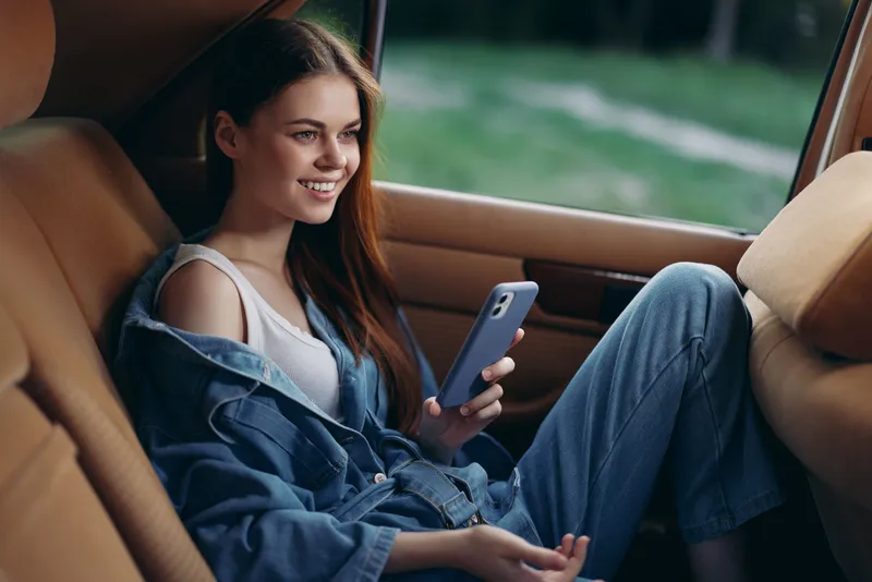 Young woman enjoying a moment in a car, wearing denim overalls and holding a phone, exuding happiness with a natural background.